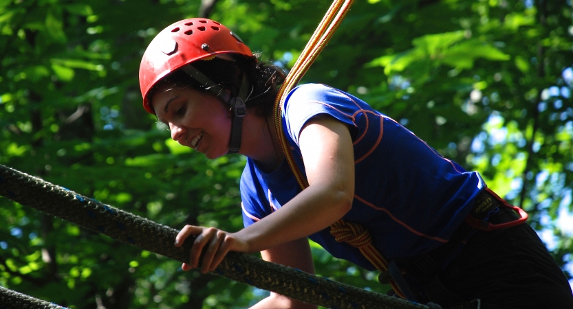 A person wearing safety gear smiles while navigating a ropes course 